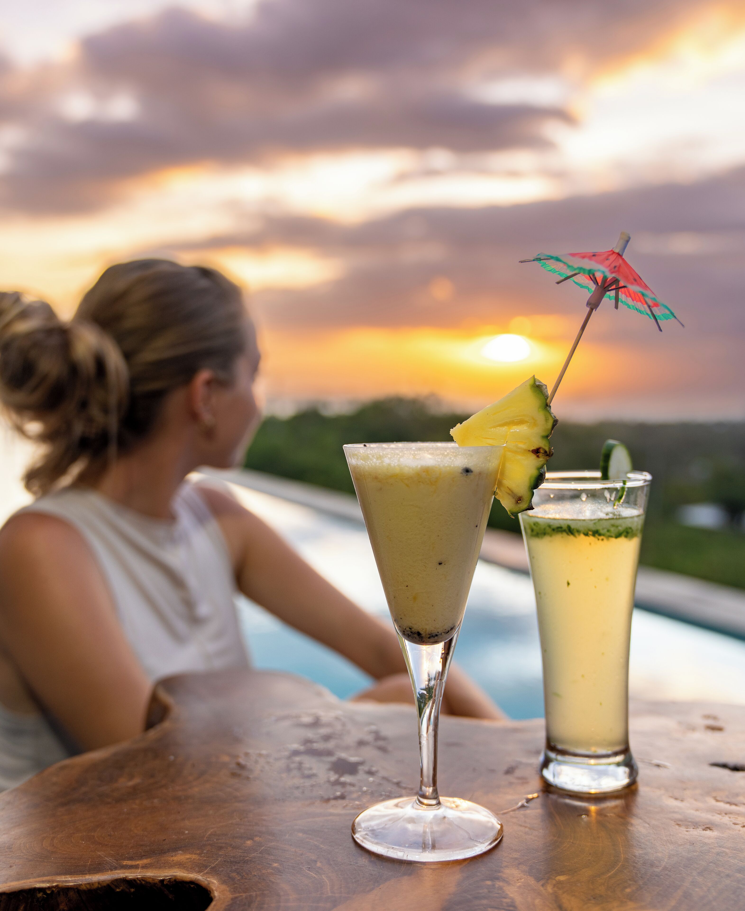 Une femme est assise au bord d’une piscine, avec deux boissons sur une table en bois.