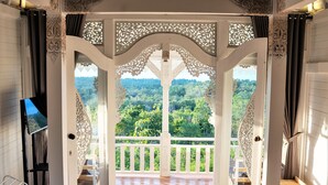 A bedroom with a large bed, a TV, and a balcony with a view of a forest. - Mambo Hill Resort (Penida Island)
