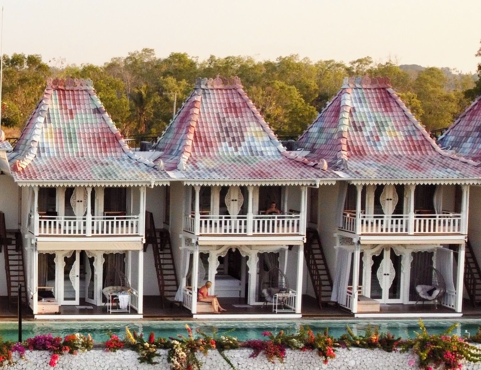 A row of three-story buildings with colorful roofs, balconies, and a pool area with flowers.