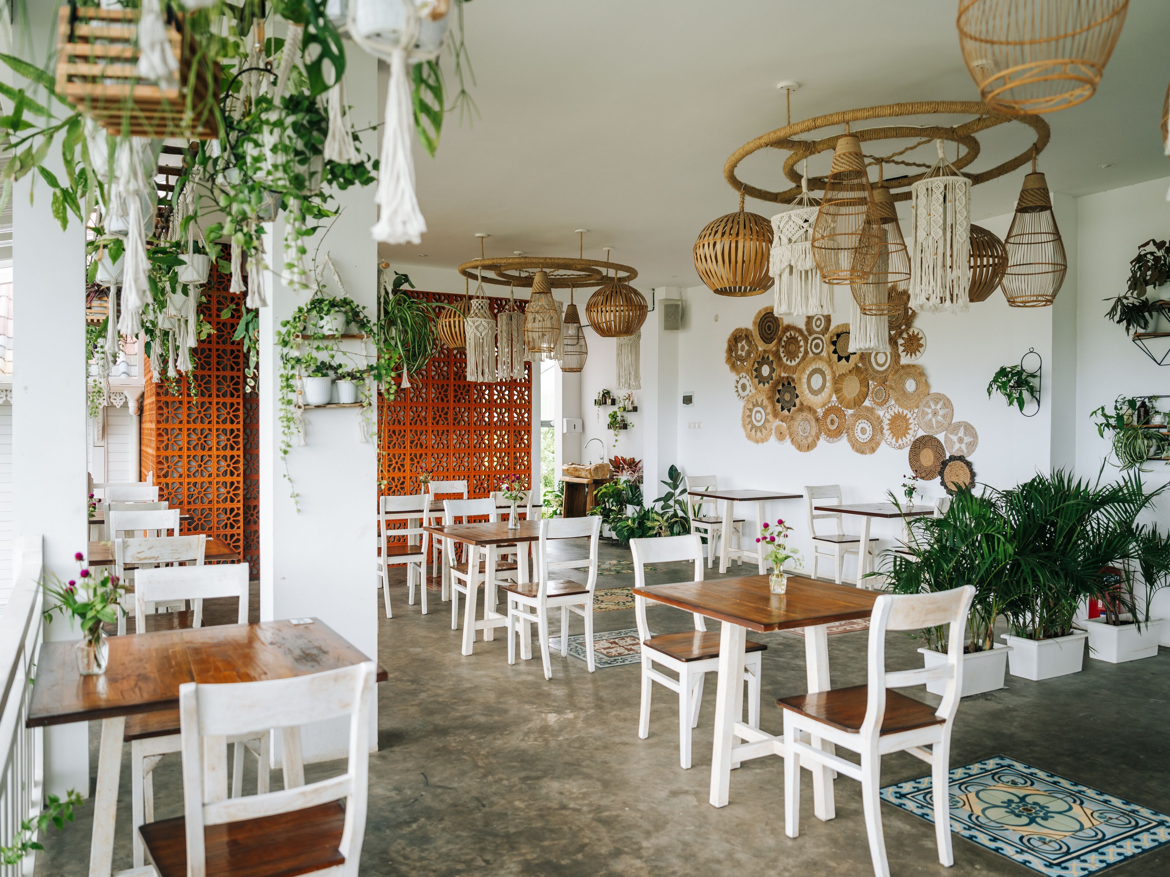 Un restaurant avec des tables et des chaises en bois peintes en blanc, des lampes en osier suspendues et une variété de plantes.
