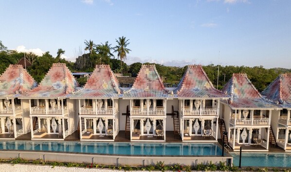 A row of white, two-story buildings with colorful roofs, a swimming pool, and palm trees in the background.