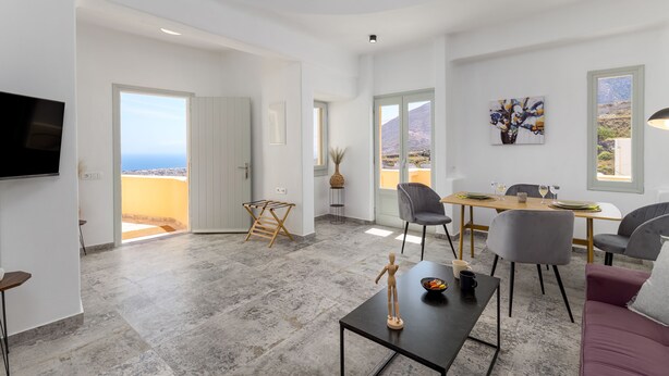A modern living room with a dining area, large windows, and a view of the landscape. - Geminos Complex Villas - Standard Villa (Santorini)