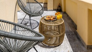 A balcony with wicker chairs, a round table with food and drinks, and potted plants. - Geminos Complex Villas - Standard Villa (Santorini)
