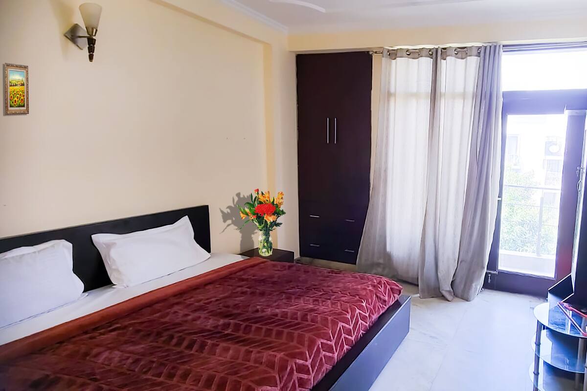 Une chambre à coucher comprenant un lit, une table de chevet, un téléviseur, une armoire et une fenêtre avec des rideaux.