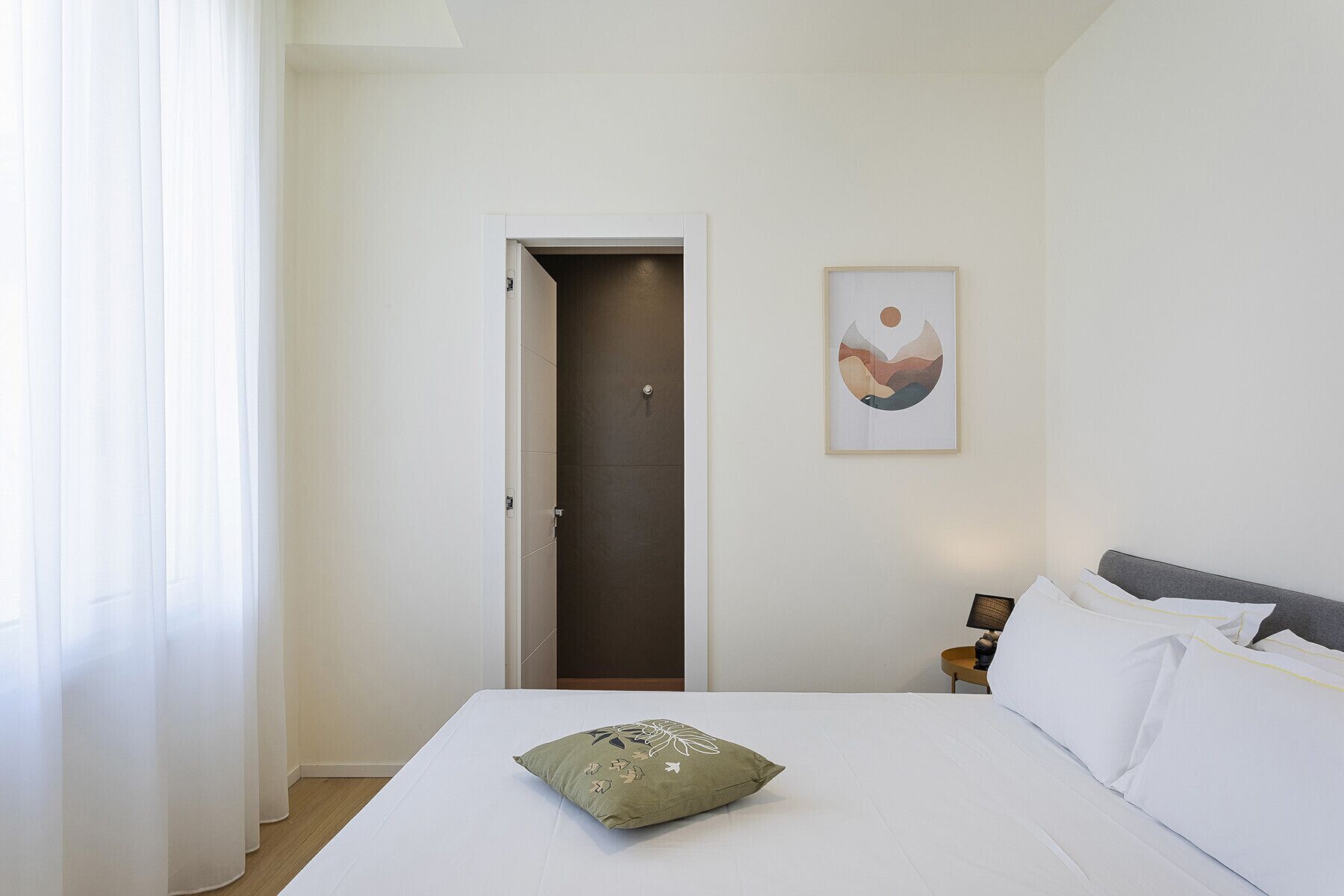A hotel room with a bed, a pillow, a door, and a framed picture on the wall.