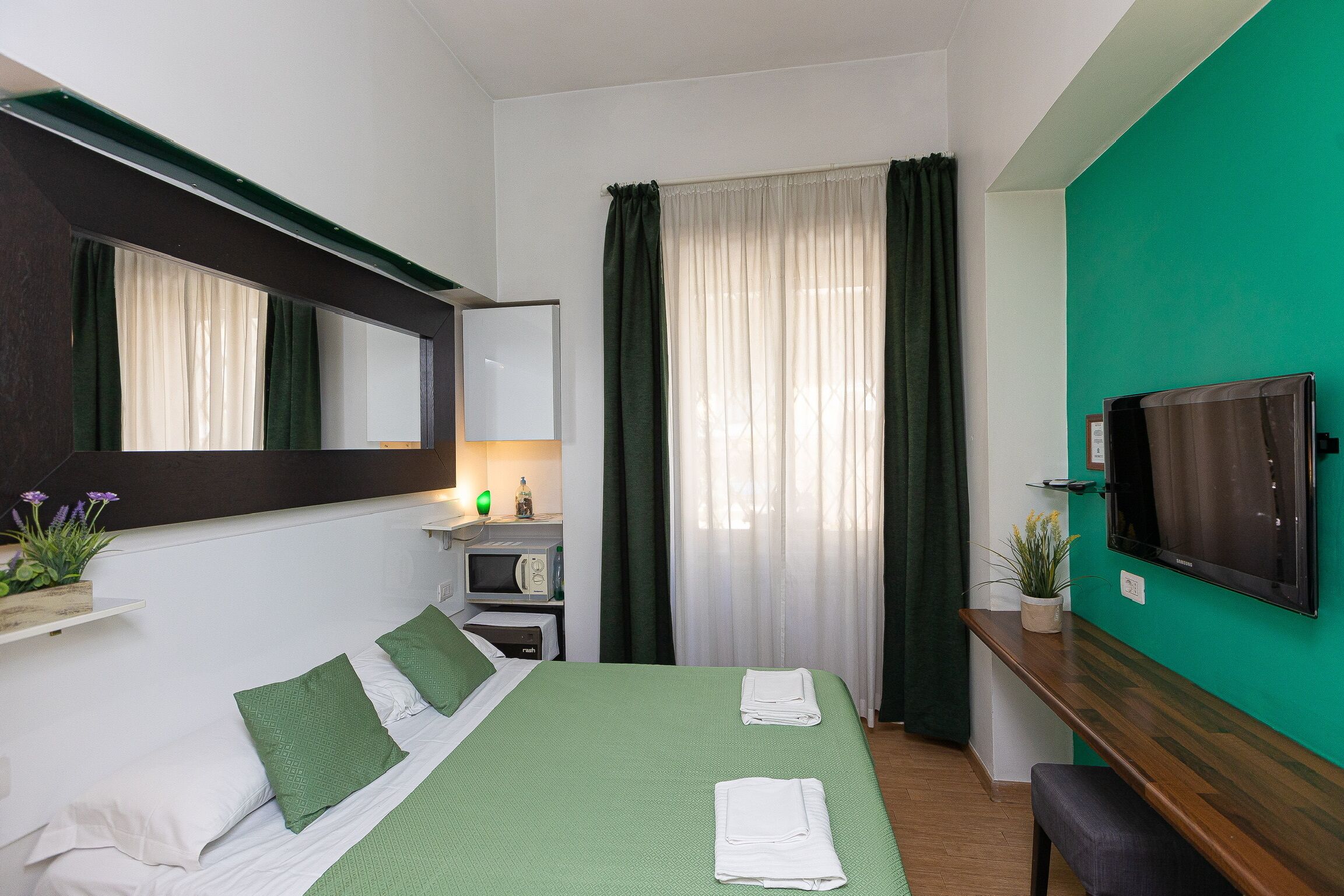 A hotel room with a bed, a TV, a desk, and a window with curtains.