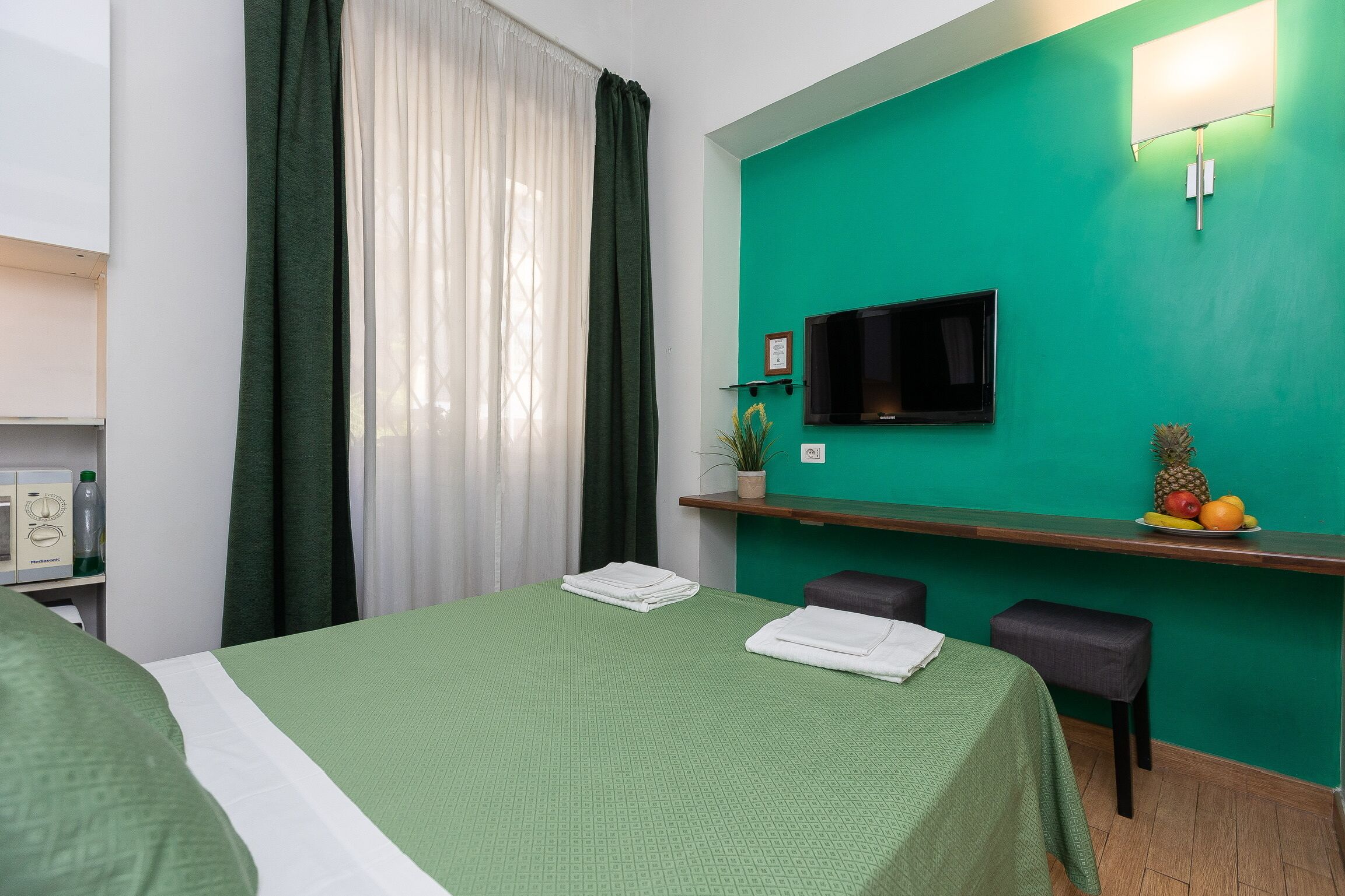 A hotel room with a green bedspread, a flat-screen TV mounted on the wall, a small shelf with fruits, and a window with green curtains.