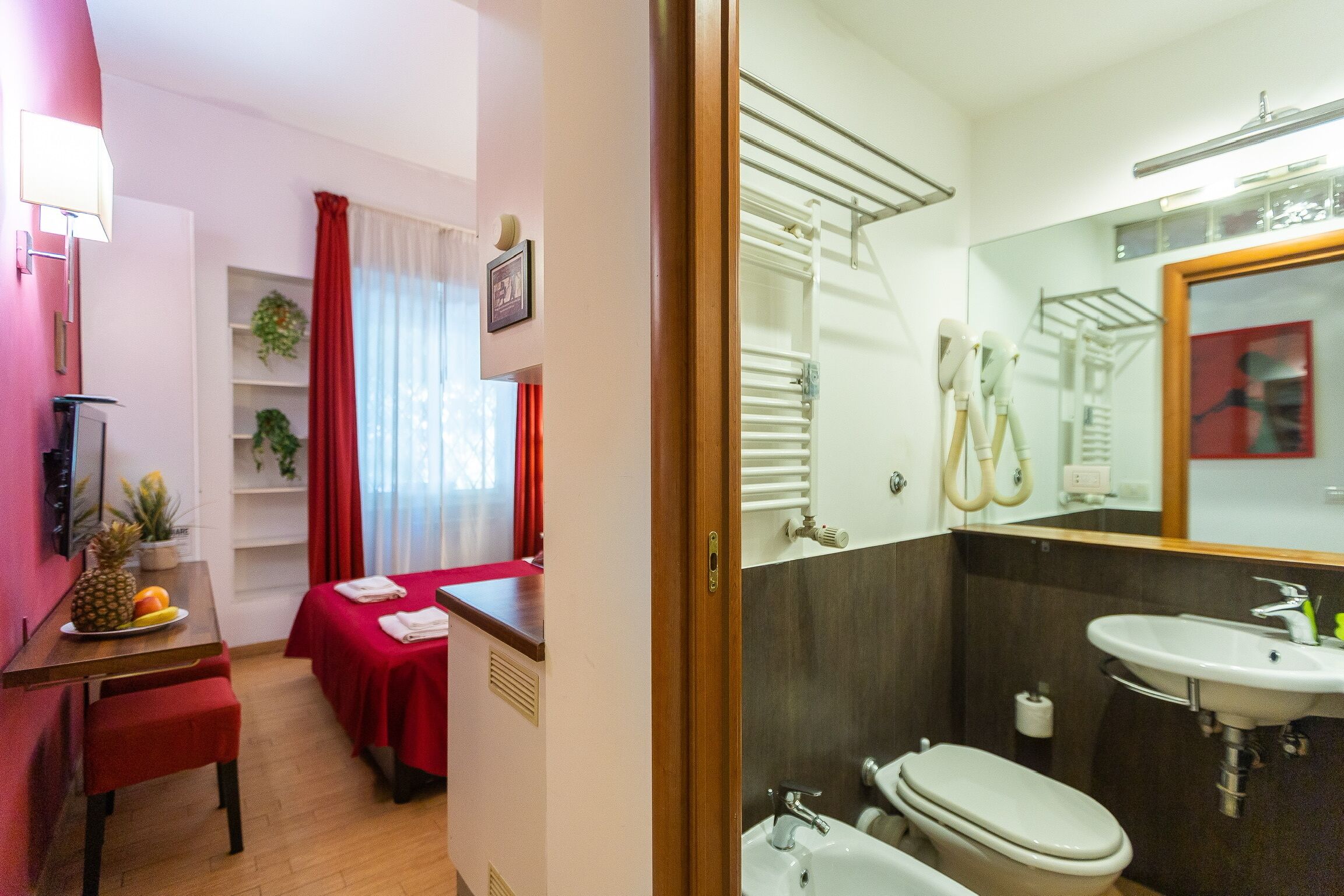 Habitación de hotel con comedor, cama con ropa de cama roja, baño con lavamanos y retrete, y un espejo que refleja la habitación.