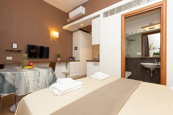 A hotel room with a bed, a table with a fruit bowl, a TV, a wall-mounted light, and a bathroom visible through an open door. - InternoUno (Rome)
