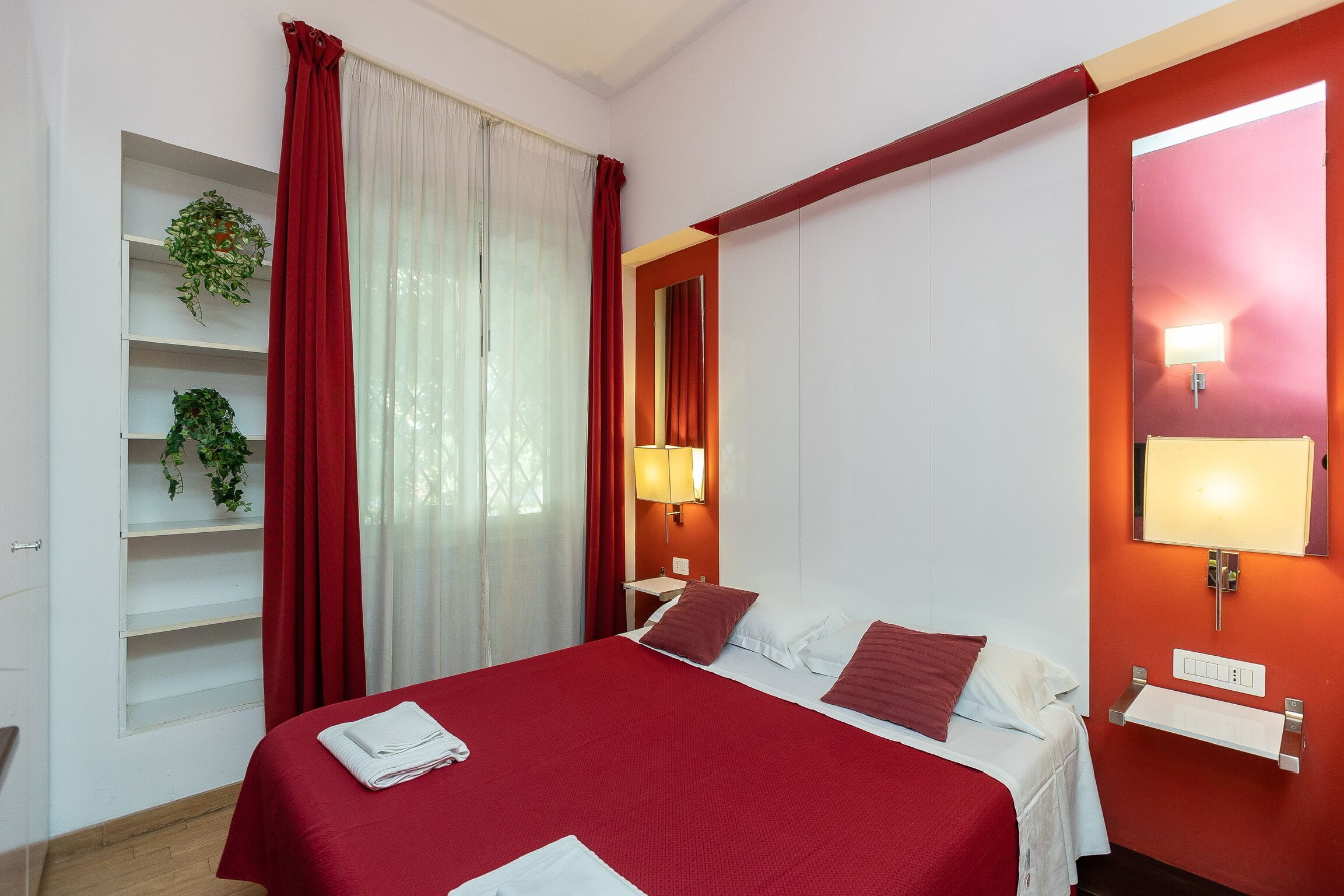 A hotel room with a bed, red curtains, a bedside lamp, a shelf with plants, and a red wall with a mirror and light fixture.