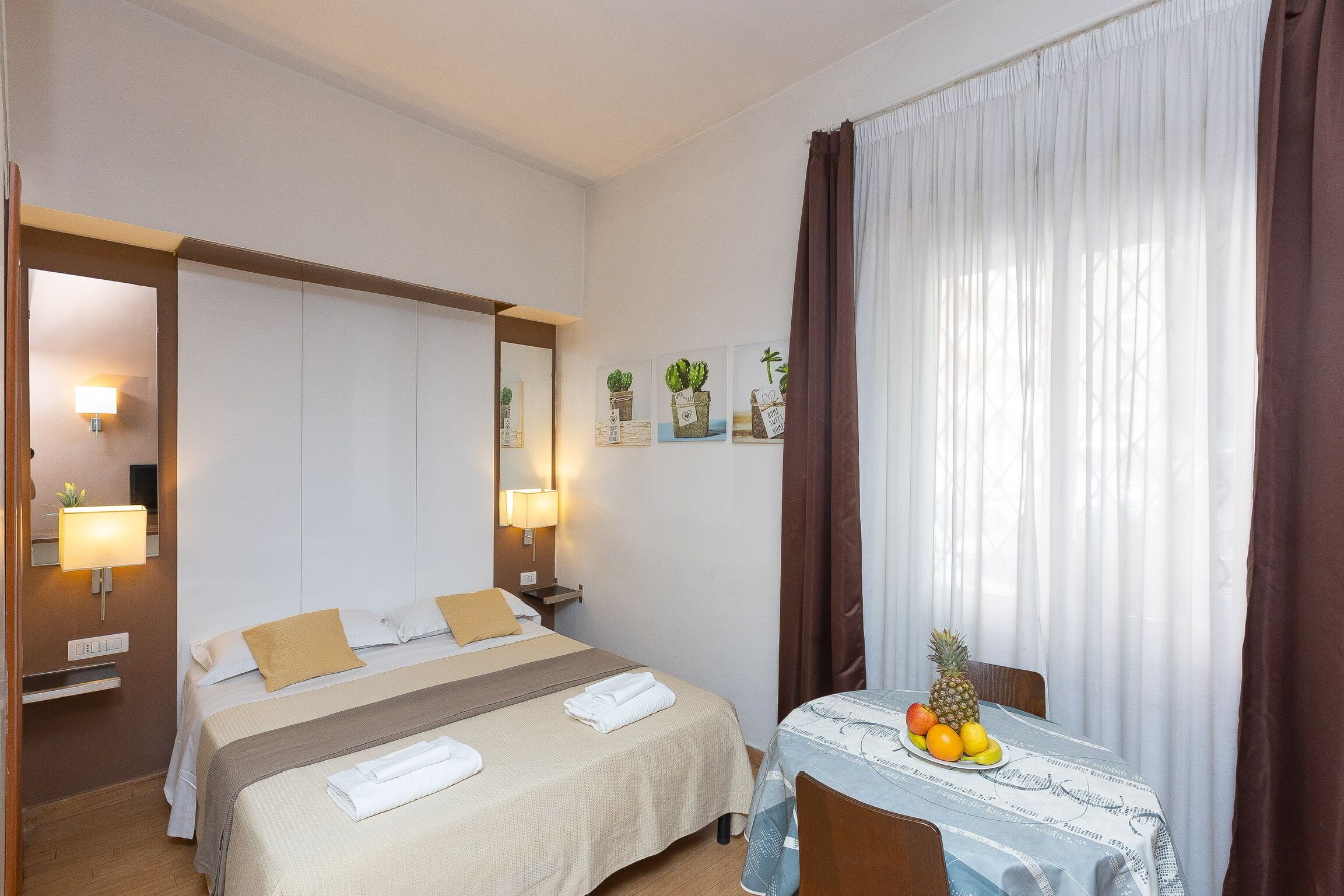 Una habitación de hotel con cama, mesitas de noche, una pequeña mesa con fruta y una ventana con cortinas.
