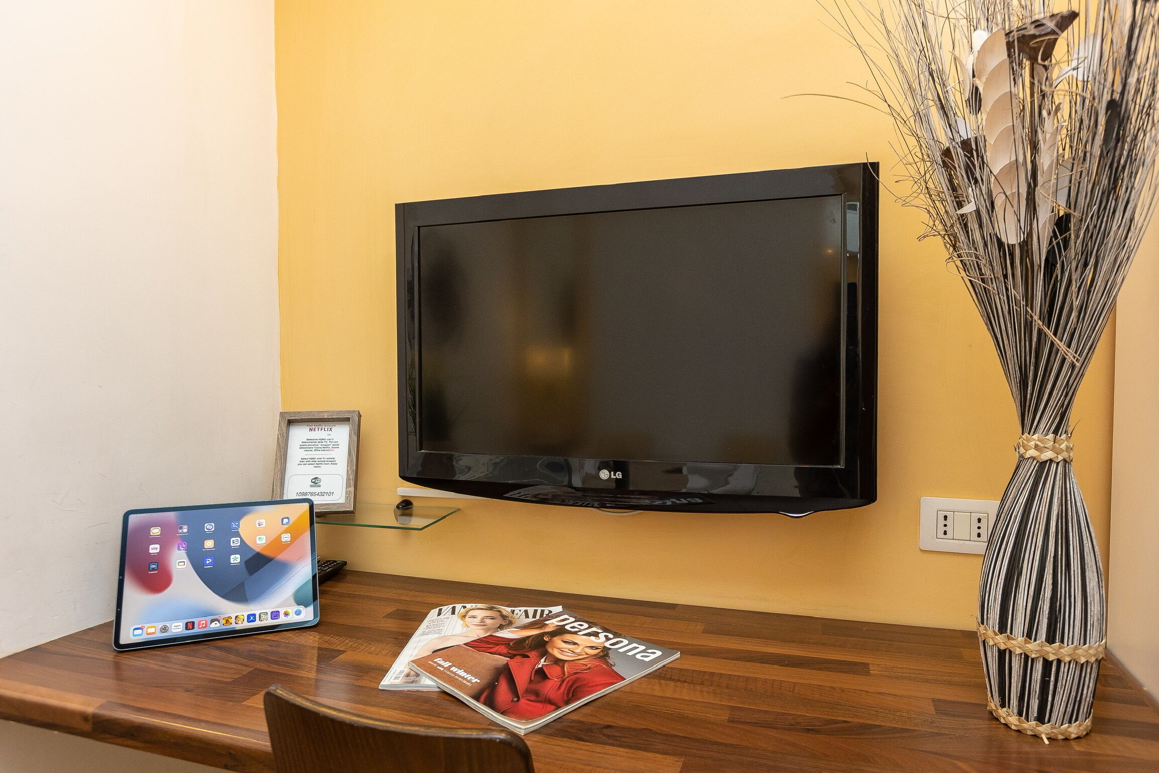 A flat-screen TV mounted on a yellow wall, a tablet on a wooden table, and a magazine on the table.