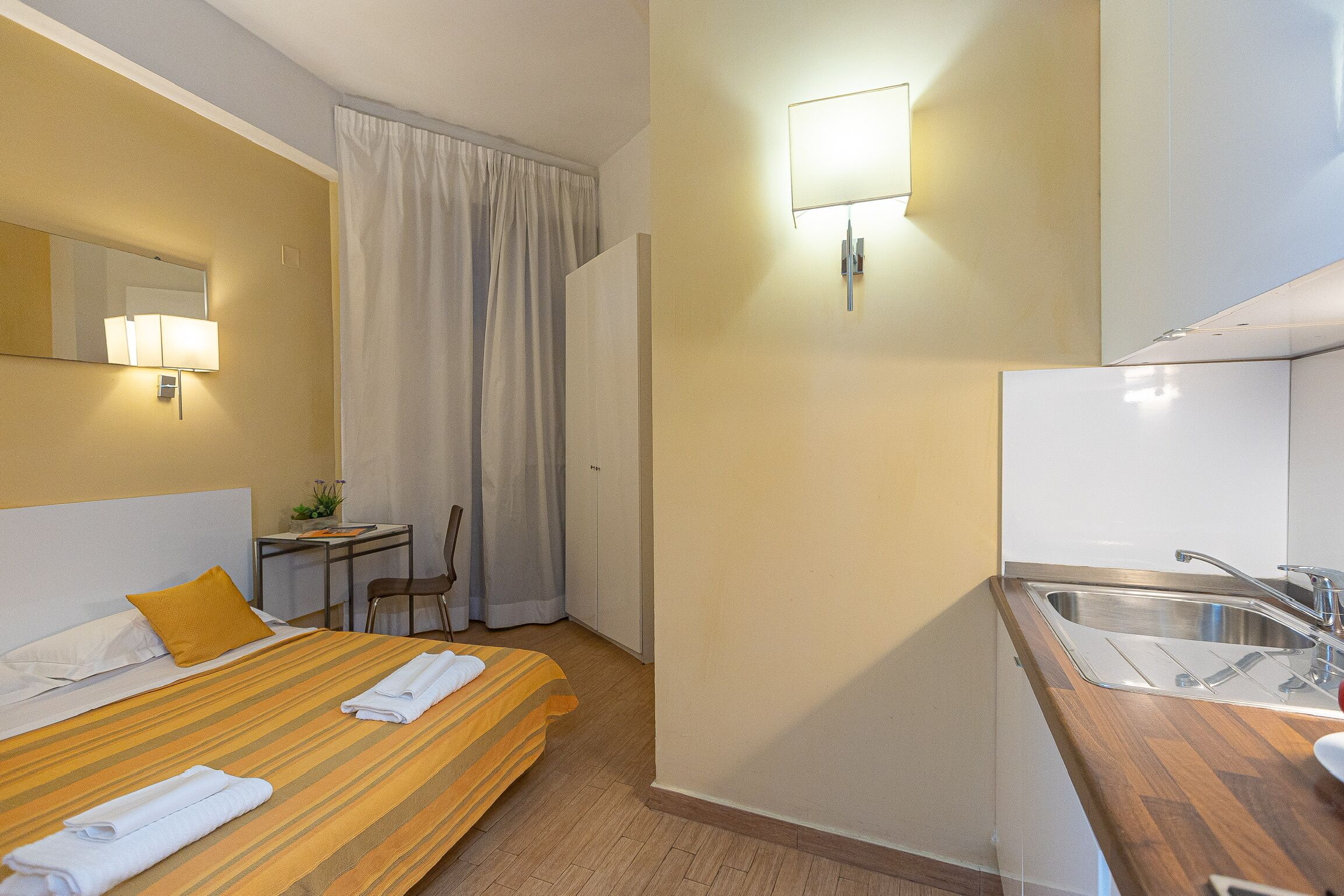 A hotel room with a bed, a desk, a chair, a small table with a plant, a window with curtains, and a kitchenette with a sink.