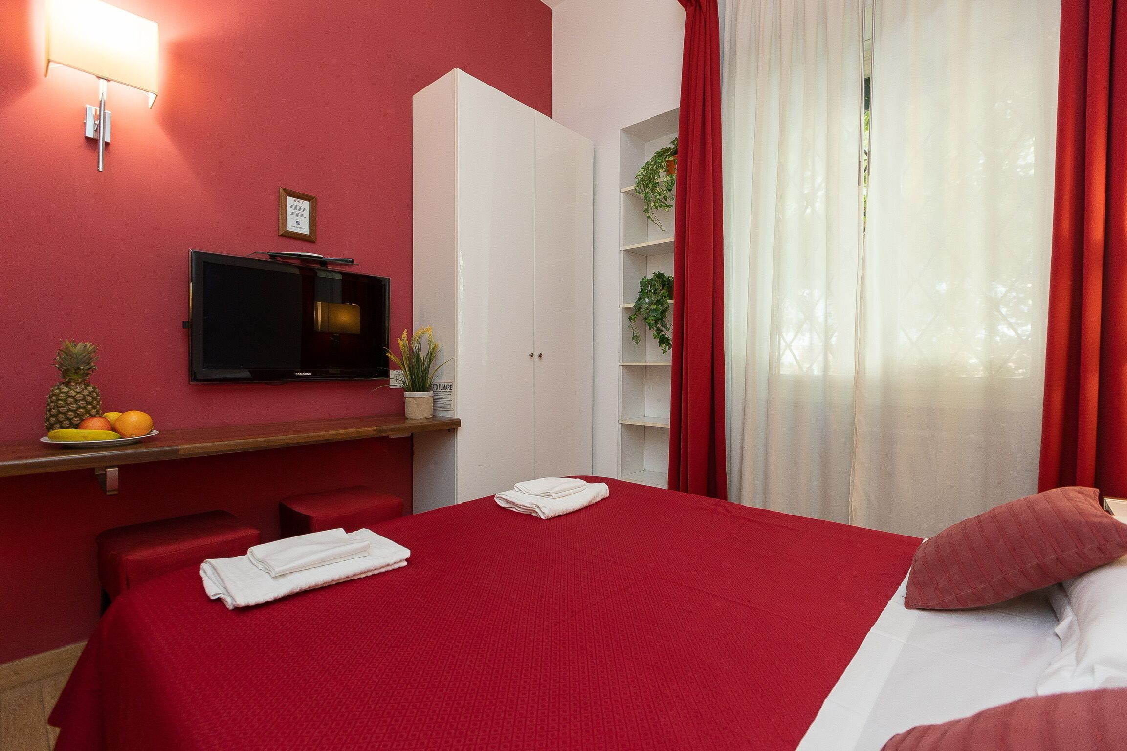 Una habitación de hotel con una colcha roja, un televisor de pantalla plana montado en la pared, una planta pequeña sobre la mesita de noche y una ventana con cortinas rojas.