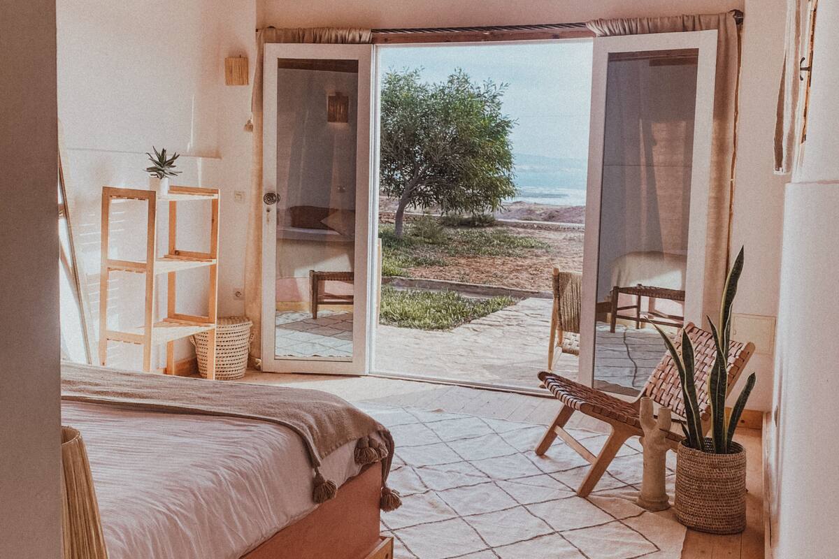 A bedroom with a bed, a wooden floor, a view of the beach, and a sliding glass door leading to an outdoor seating area.