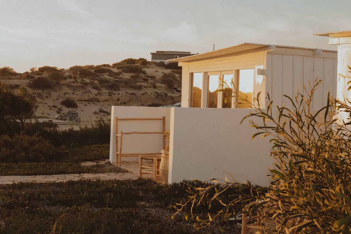 A modern, minimalist house with a large glass window, situated on a hillside with sparse vegetation.