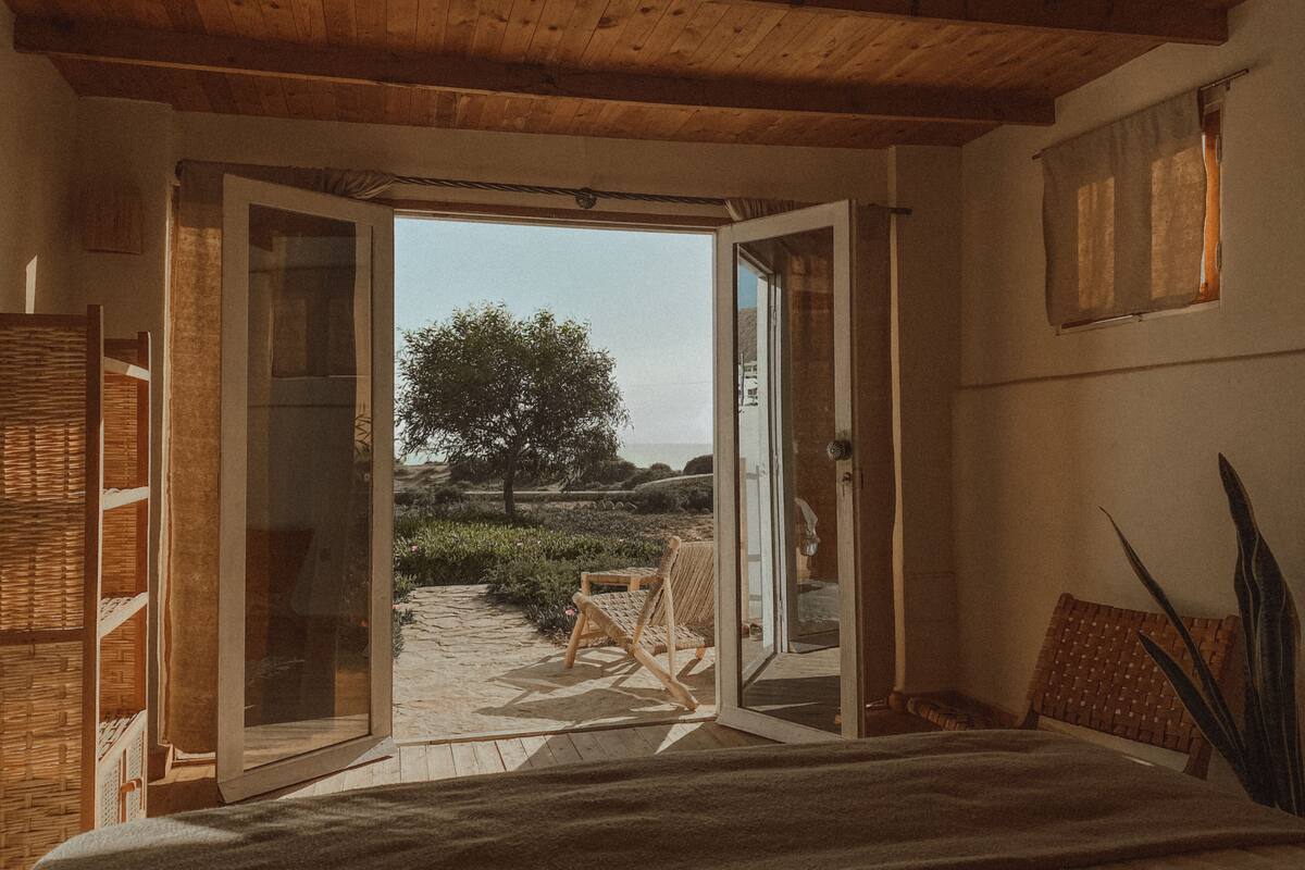 An open doorway leading to a garden with a tree, a wicker chair, and a bed with a view.
