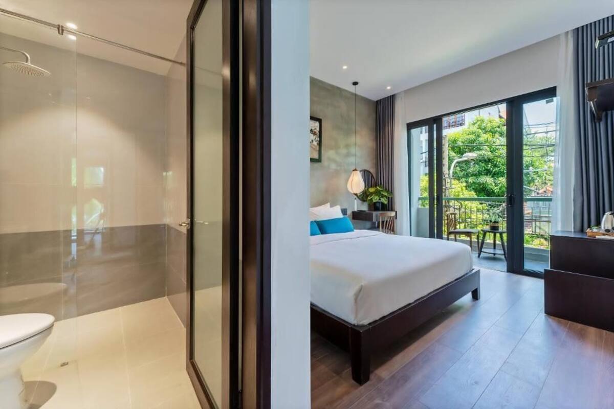 A modern hotel room with a glass shower enclosure, a large bed with a view of the outdoors, and a balcony with a dining area.