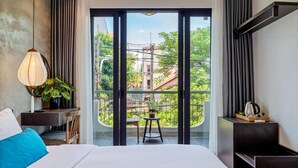 A hotel room with a large bed, a desk with a lamp, a small table with a plant, and a balcony with a view of greenery and buildings. - Lacasa Old Town Villa (Da Nang)