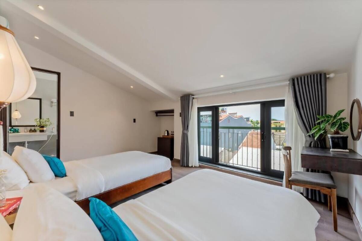 A hotel room with two beds, a desk, a chair, a large window with a view of buildings, and a balcony.