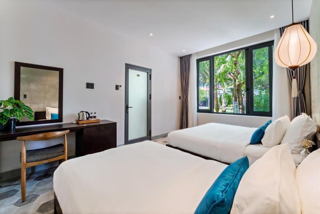 A modern hotel room with two beds, a desk, and a large window offering a view of greenery.