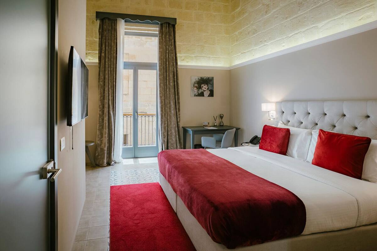 A hotel room with a large bed, a desk, a chair, a TV, and a balcony with curtains.