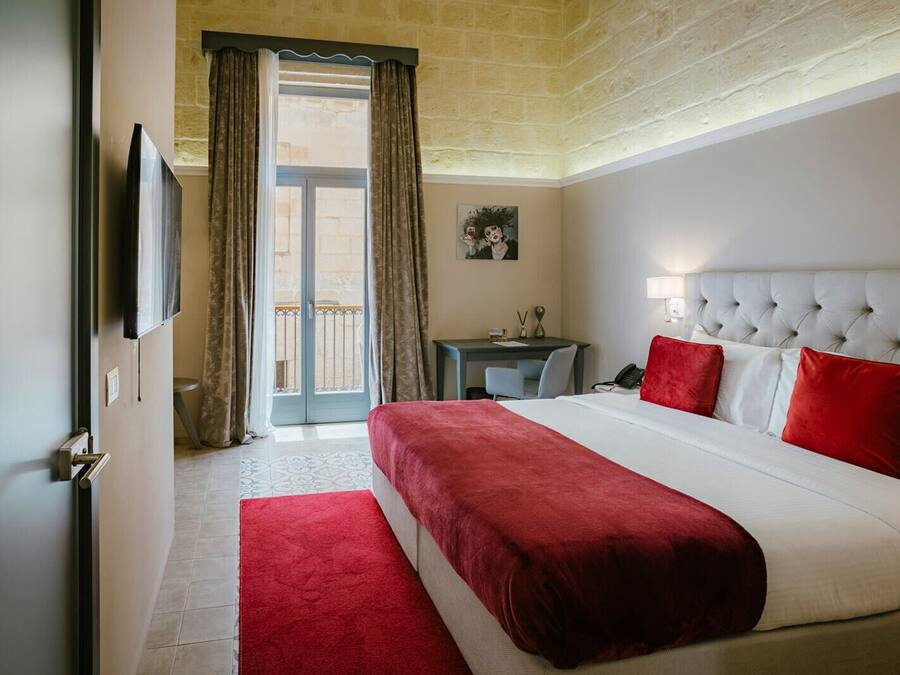 A hotel room with a large bed, a desk, a chair, a TV, and a balcony with curtains.