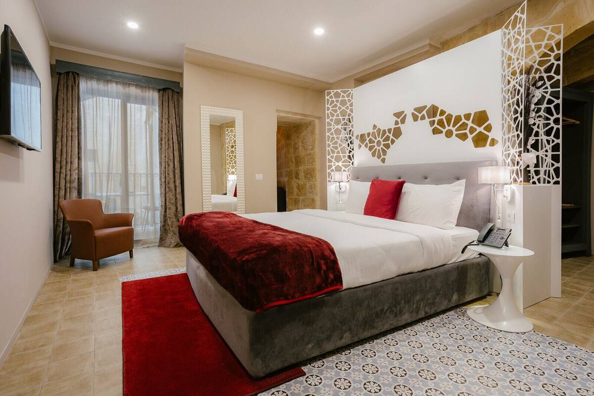 A modern hotel bedroom with a large bed, a red throw, a grey headboard, and a white nightstand with a phone.