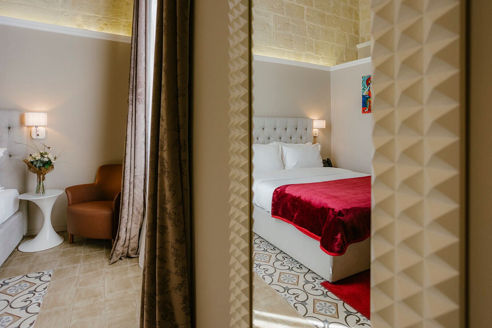 Habitación de hotel con cama, una silla, una mesita con un florero y una lámpara.