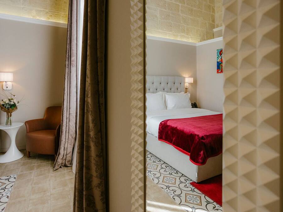 Habitación de hotel con cama, una silla, una mesita con un florero y una lámpara.