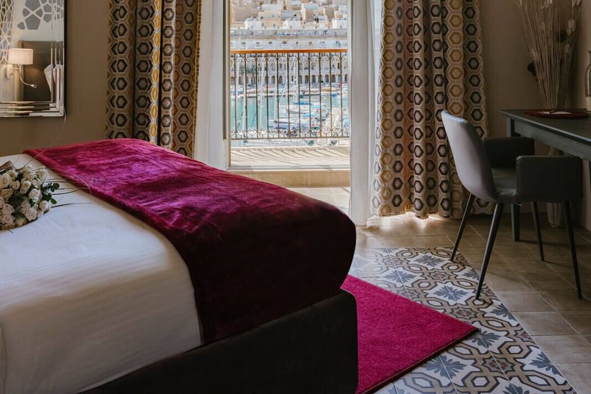 A hotel room with a bed, a desk, and a view of a cityscape through large windows.