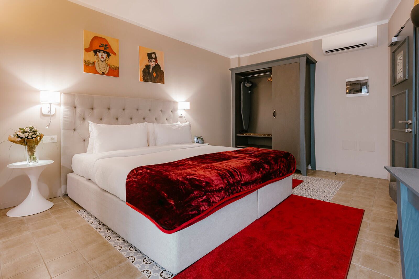 Un dormitorio moderno con una cabecera capitonada, una cama blanca, un plaid rojo, una mesita de noche con un jarrón de flores y una alfombra roja.