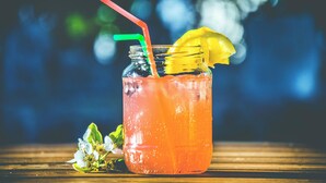 Een glas met een rode rietje en een gele schijfje garnering, geplaatst op een houten oppervlak met onscherpe begroeiing op de achtergrond.