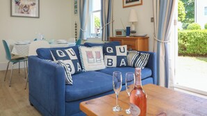 A living room with a blue sofa, two wine glasses, a bottle of wine, and a dining area with a table and chairs. - 5 Combehaven (Salcombe)