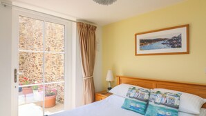 A bedroom with a bed, a nightstand, a lamp, a framed picture on the wall, and a view of a stone wall and potted plants through a sliding glass door. - 5 Combehaven (Salcombe)