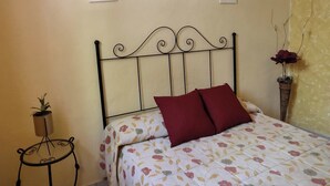 Un llit amb una col·cha floral i dos coixins vermells, una taula rodona petita amb una planta, i tres quadres a la paret.