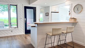 A modern kitchen with a bar area, white wooden walls, and a view of the outdoors.