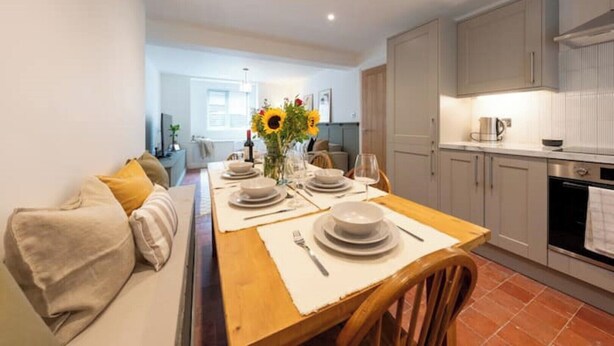 Et spiseområde med et træbord dækket til fire personer, en sofa med et beige tæppe, og et køkken med grå skabe og et flisebeklædt backsplash.