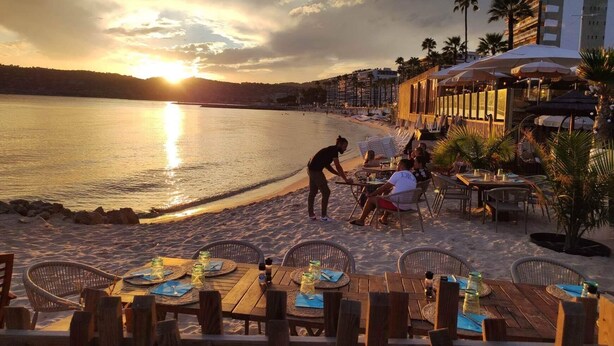 Un restaurante frente al mar con terraza al aire libre, un camarero atendiendo a los clientes y un atardecer en el fondo.