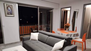 Un salón moderno con un sofá gris, cojines blancos, una mesita pequeña blanca y sillas naranjas.