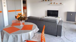 Una sala de estar moderna con un sofá gris, una mesa de comedor con detalles en naranja y un televisor de pantalla plana sobre un soporte blanco.