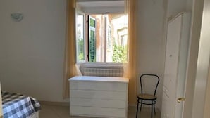 A small room with a bed, a window with curtains, a radiator, a chair, and a door.