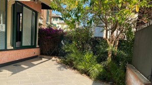 A paved courtyard with a tiled floor, surrounded by greenery and a building with a green window frame.