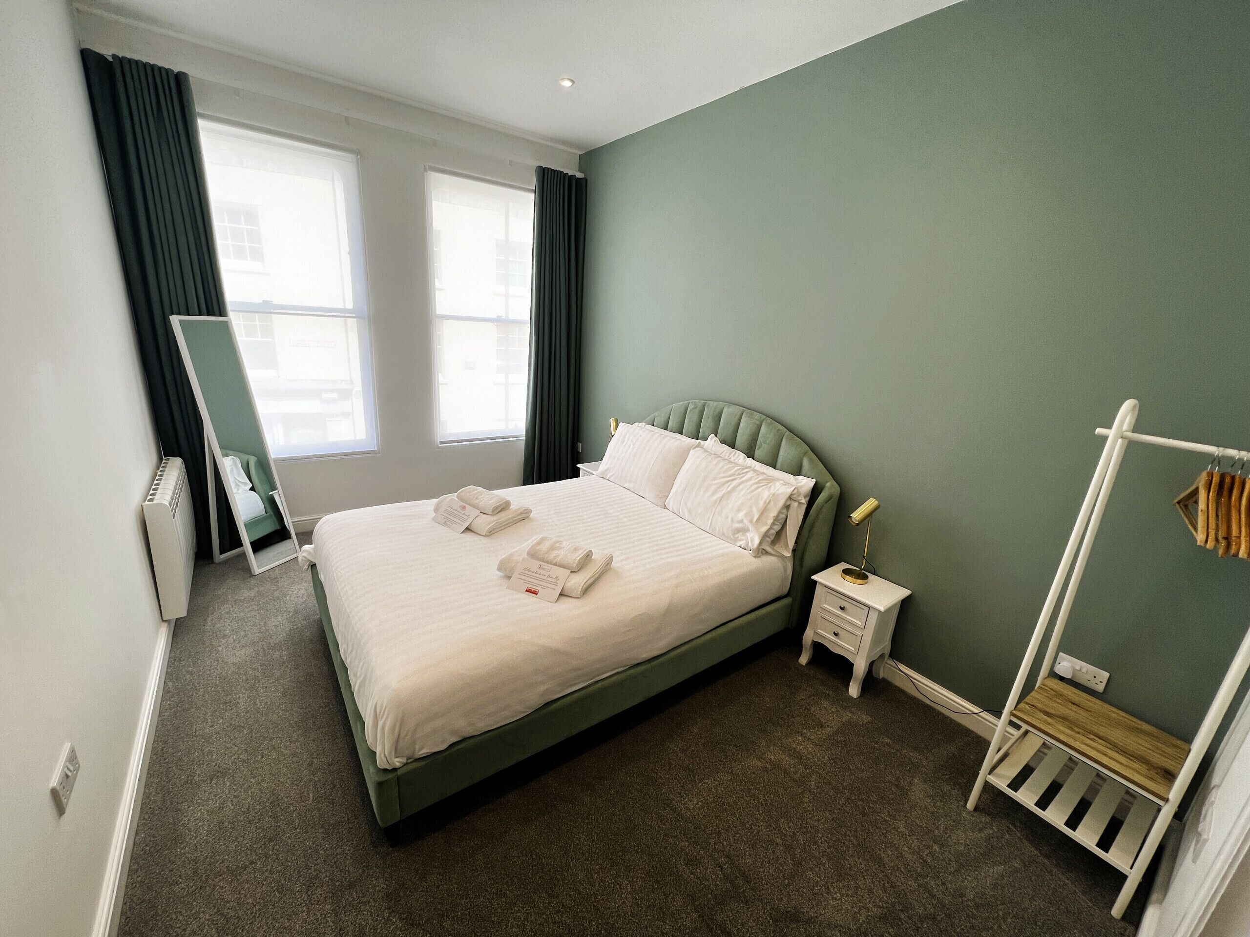 A neatly made bed with pillows and folded towels, a bedside table with a lamp, a mirror, and a radiator.