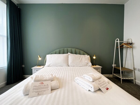 A neatly made bed with white linens and towels, a bedside lamp, and a towel rack with folded towels.