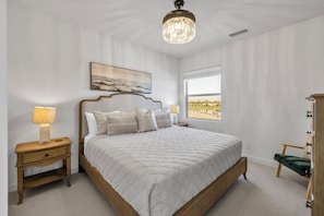 A bedroom with a bed, nightstands, a lamp, a painting, a window with a view, and a chandelier.