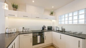 A modern kitchen with white cabinets, a black countertop, a built-in oven, and a window with blinds. - Monopoly Breeze House (Brighton and Hove)