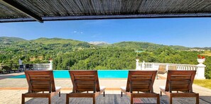 A terrace with four wooden chairs facing a pool and a mountainous landscape.