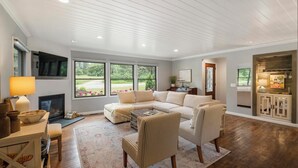 Een ruime woonkamer met een grote modulaire bank, een open haard en uitzicht op een tuin door grote ramen.