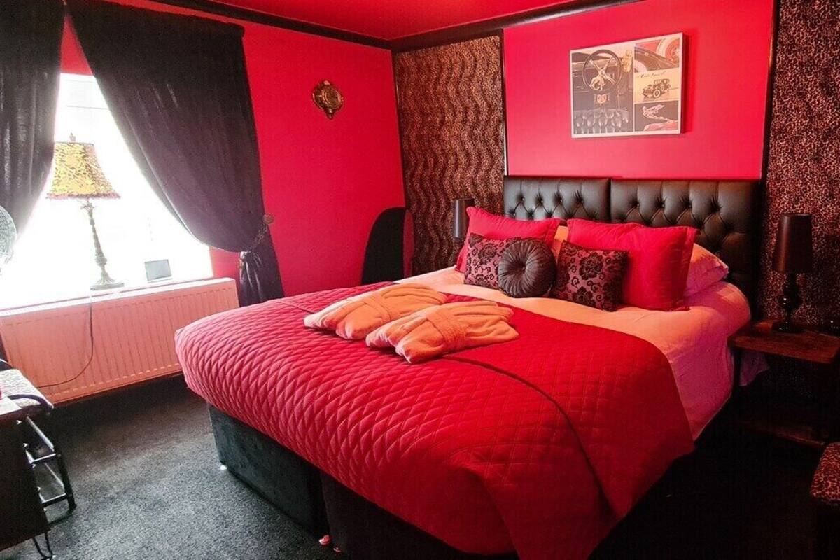 A red-themed hotel room with a large bed, a bedside table with toiletries, and a window with curtains.